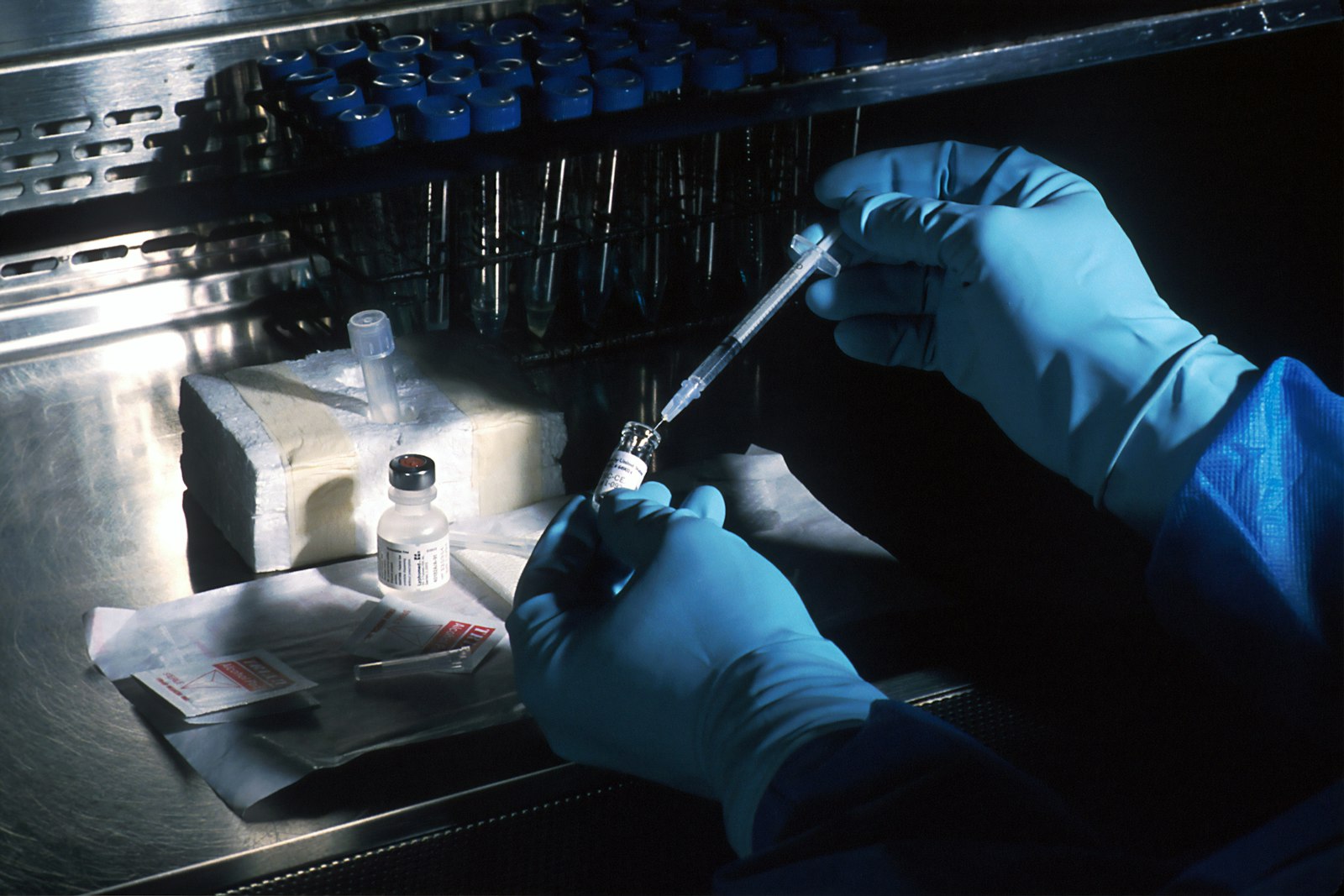 Gloved hands in a laboratory drawing clear liquid from a small glass vial with a syringe