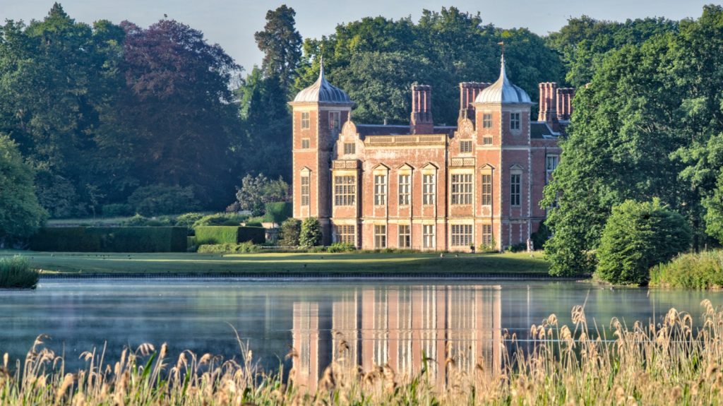 A historic English manor house reflected in a calm lake with reeds at sunrise, evoking a Jane Austen period drama