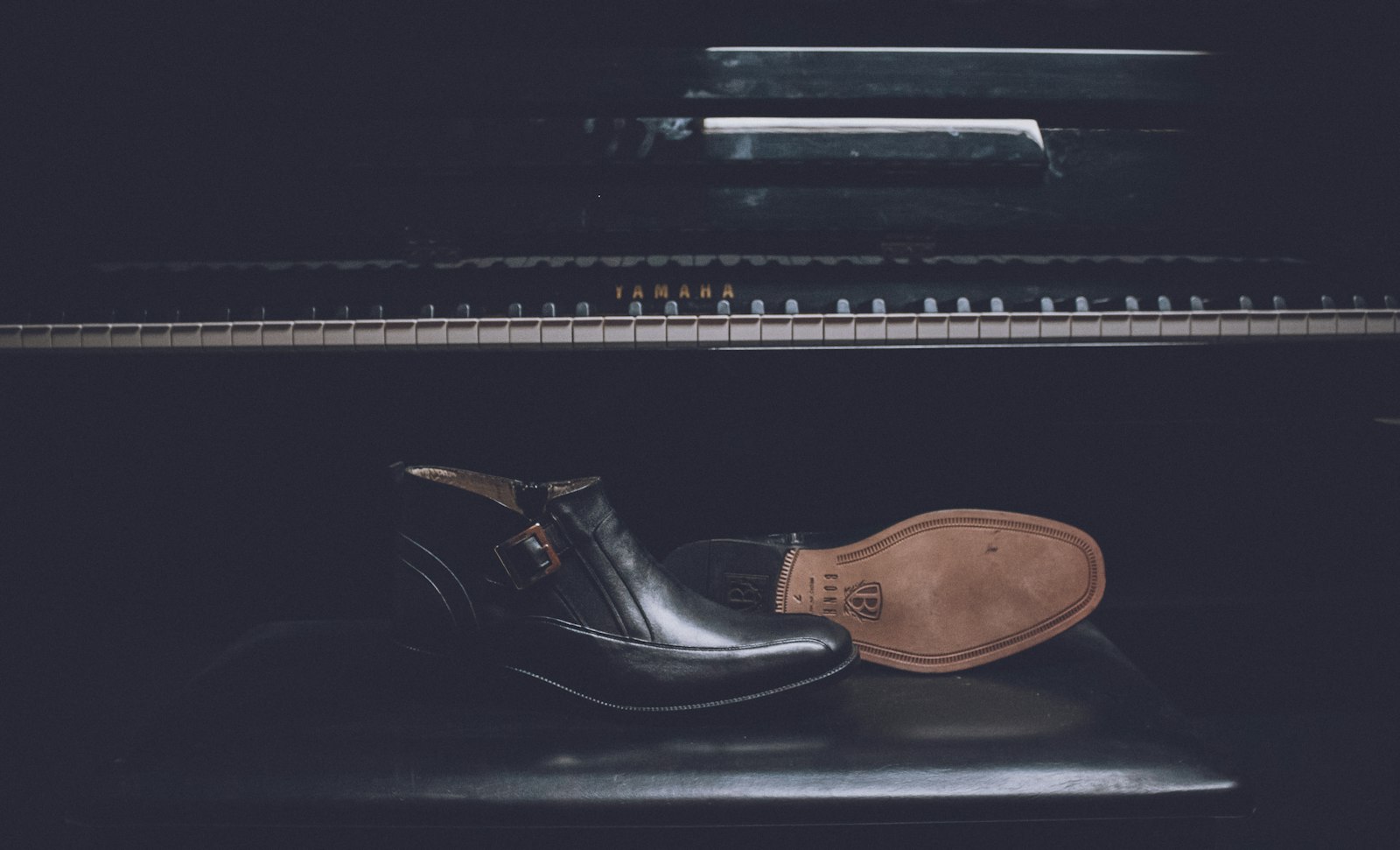 A pair of black patent leather dress shoes resting on a piano bench in low light