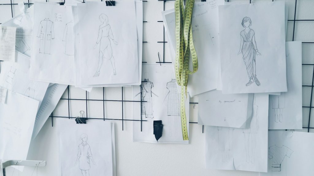 Black and white fashion sketches with garment measurements pinned on a grid board in a design studio