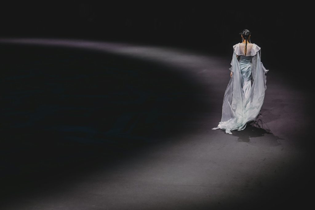 A model in a flowing light blue gown walking along a dark fashion show runway