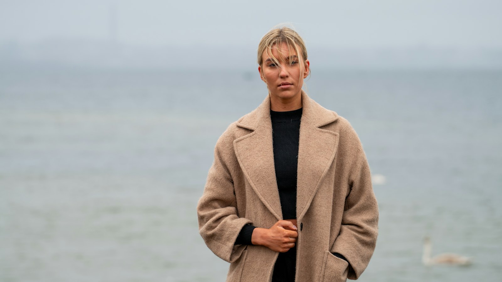 Woman wearing an elegant beige coat standing by the sea on a calm grey day