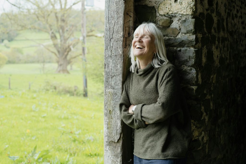 Smiling older woman leaning in a rustic doorway that overlooks a green field