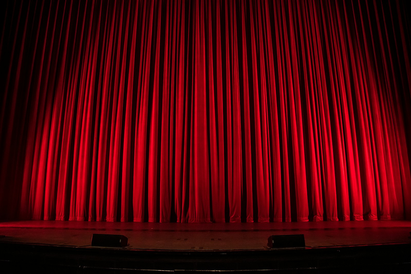 Closed red velvet theatre curtain lit by stage lights in a darkened auditorium