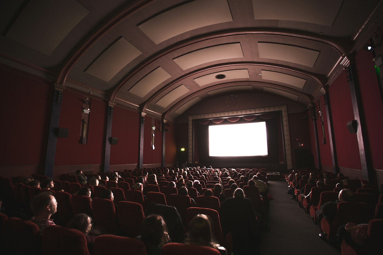 Audience seated in red velvet seats in a dark cinema facing a bright screen